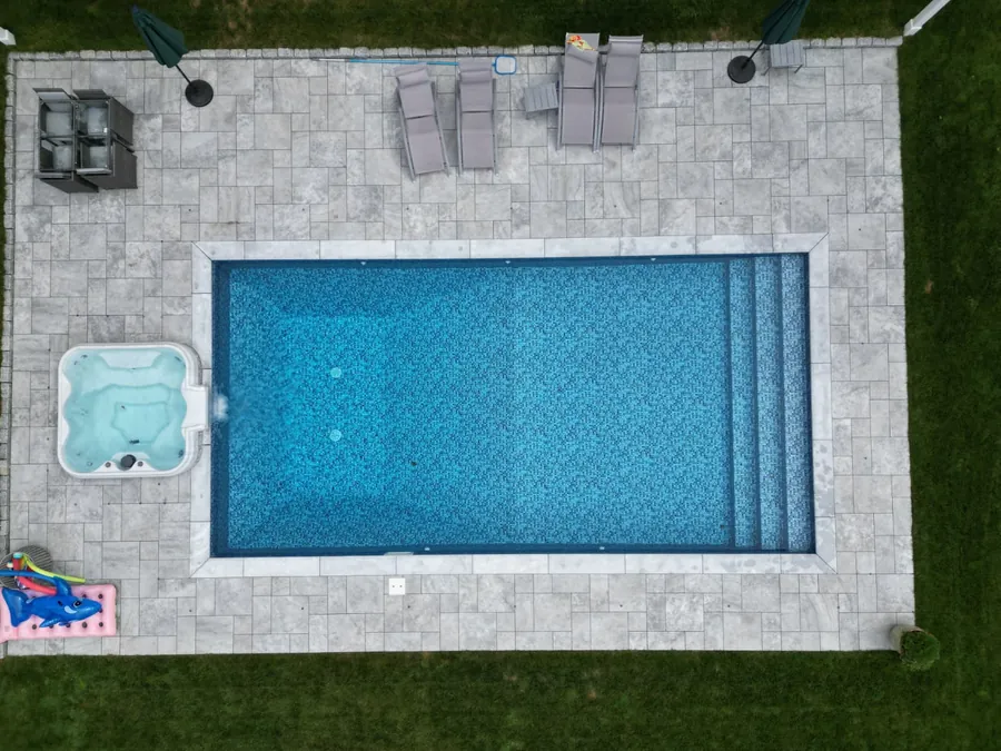 Aerial view of a rectangular pool with a hot tub on a paver patio and lawn