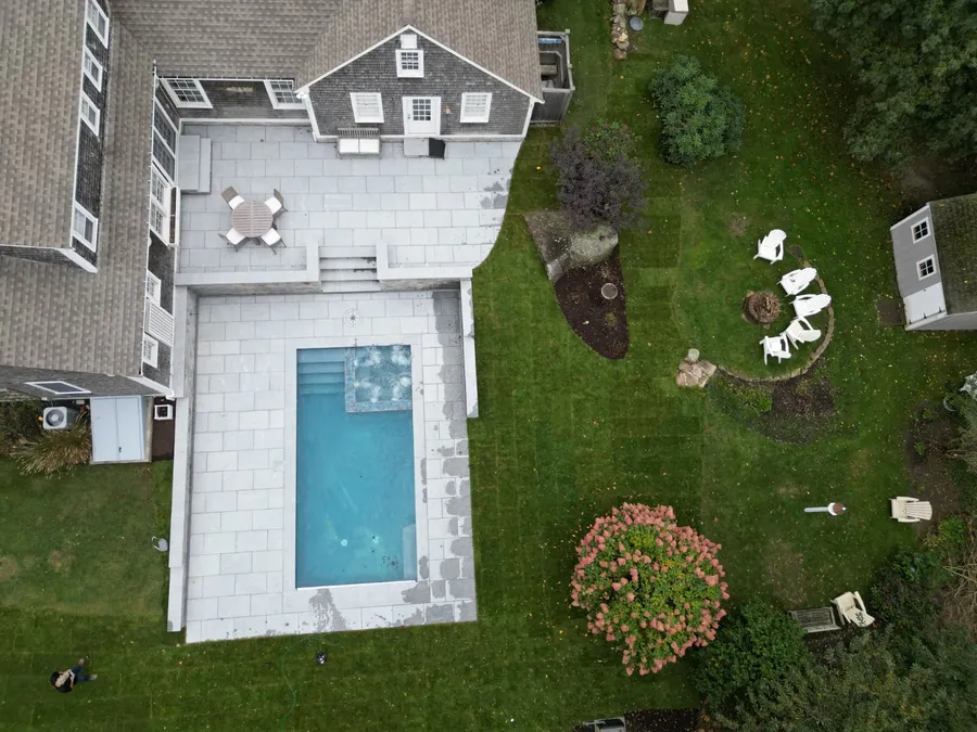 Aerial view of a rectangular pool with an in-pool spa on a stone patio behind a shingled Cape house in Barnstable, MA
