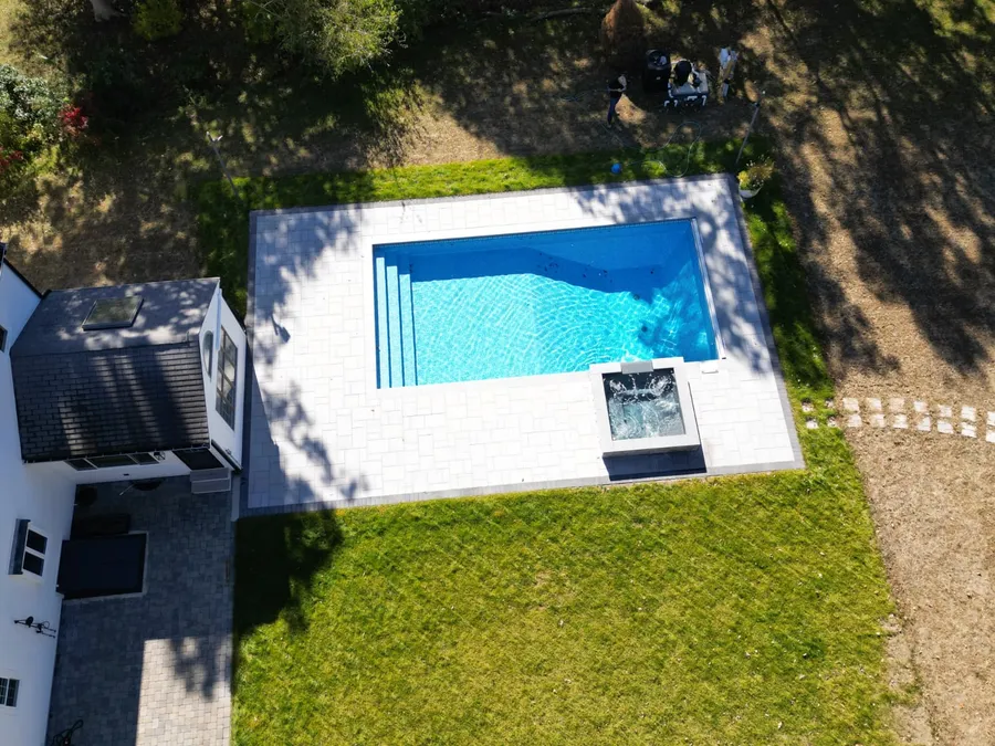 Aerial view of a new rectangular pool in Sandwich, MA with a raised spillover spa, grey paver deck and fresh lawn